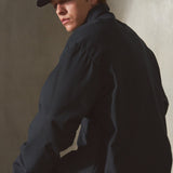 A man wearing a navy baseball cap, navy jacket, black pants, and black gloves sits on a concrete block against a textured concrete wall.