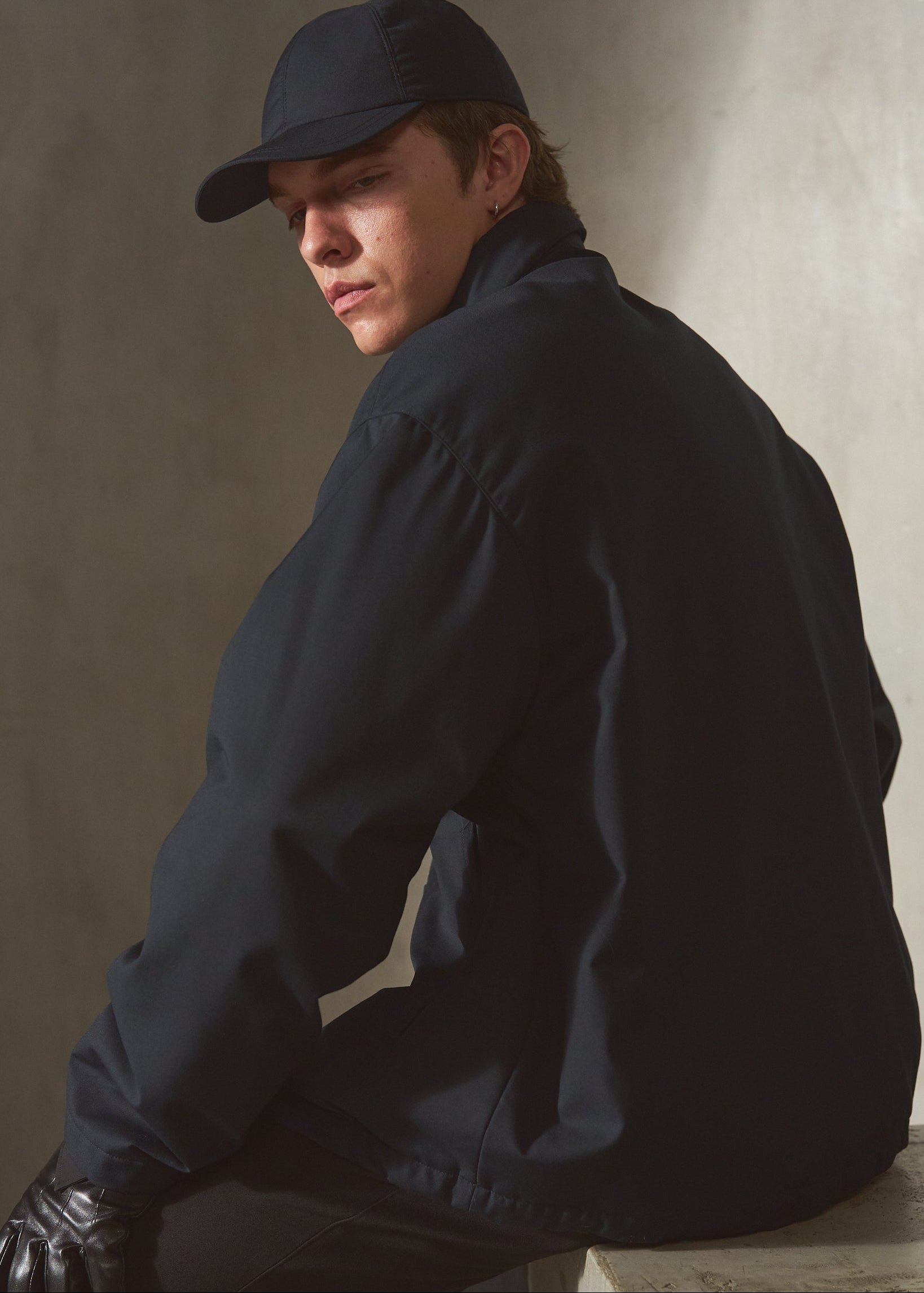 A man wearing a navy baseball cap, navy jacket, black pants, and black gloves sits on a concrete block against a textured concrete wall.