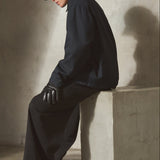 A model wearing a navy cap, navy jacket, black wide-leg trousers, and black gloves sits on a concrete block in a minimalist studio setting with a textured beige wall and floor.
