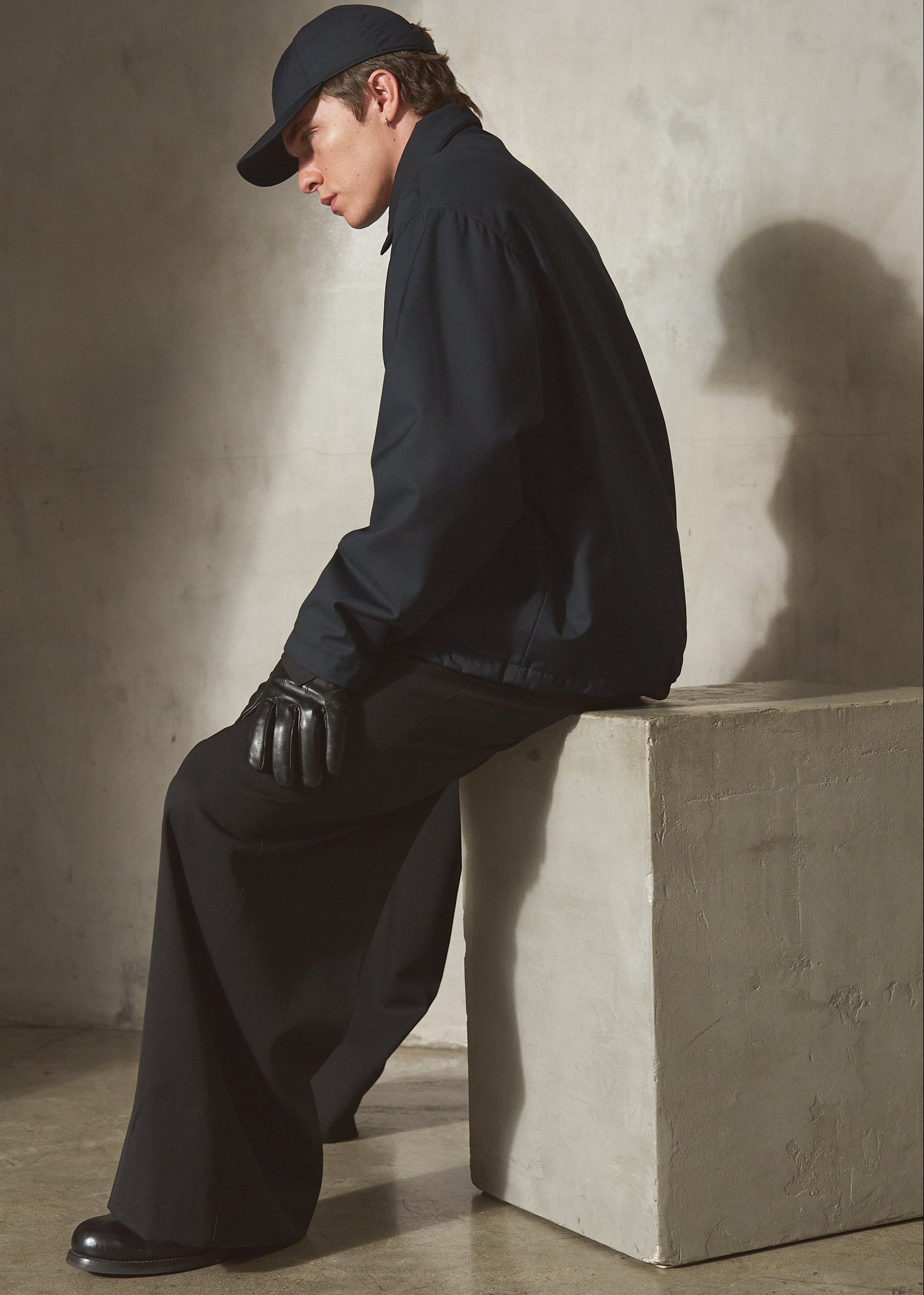 A model wearing a navy cap, navy jacket, black wide-leg trousers, and black gloves sits on a concrete block in a minimalist studio setting with a textured beige wall and floor.