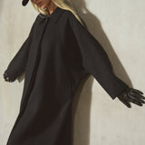 A model wearing a long black batwing coat, black gloves, and a black baseball cap leans against a textured light gray wall, casting a strong shadow.