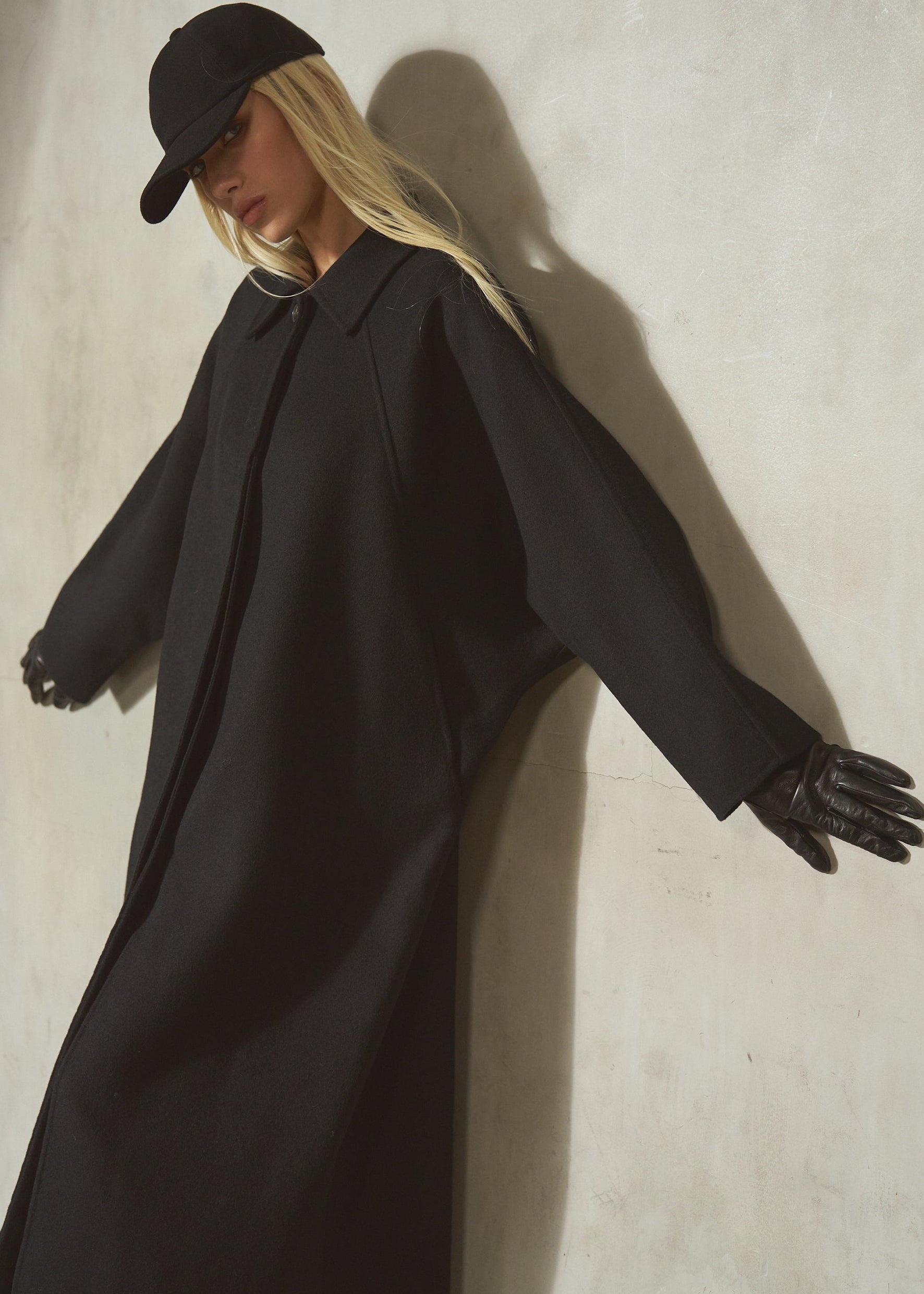 A model wearing a long black batwing coat, black gloves, and a black baseball cap leans against a textured light gray wall, casting a strong shadow.