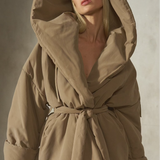 A model wearing a beige oversized puffer jacket with a large attached hood, tie belt, and cuffed sleeves, styled with a black cap, black gloves, and sheer black tights against a textured concrete wall.