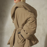A model wearing a voluminous beige puffer coat with a matching waist tie belt, paired with a black baseball cap and sheer black tights, posing against a textured gray concrete wall.