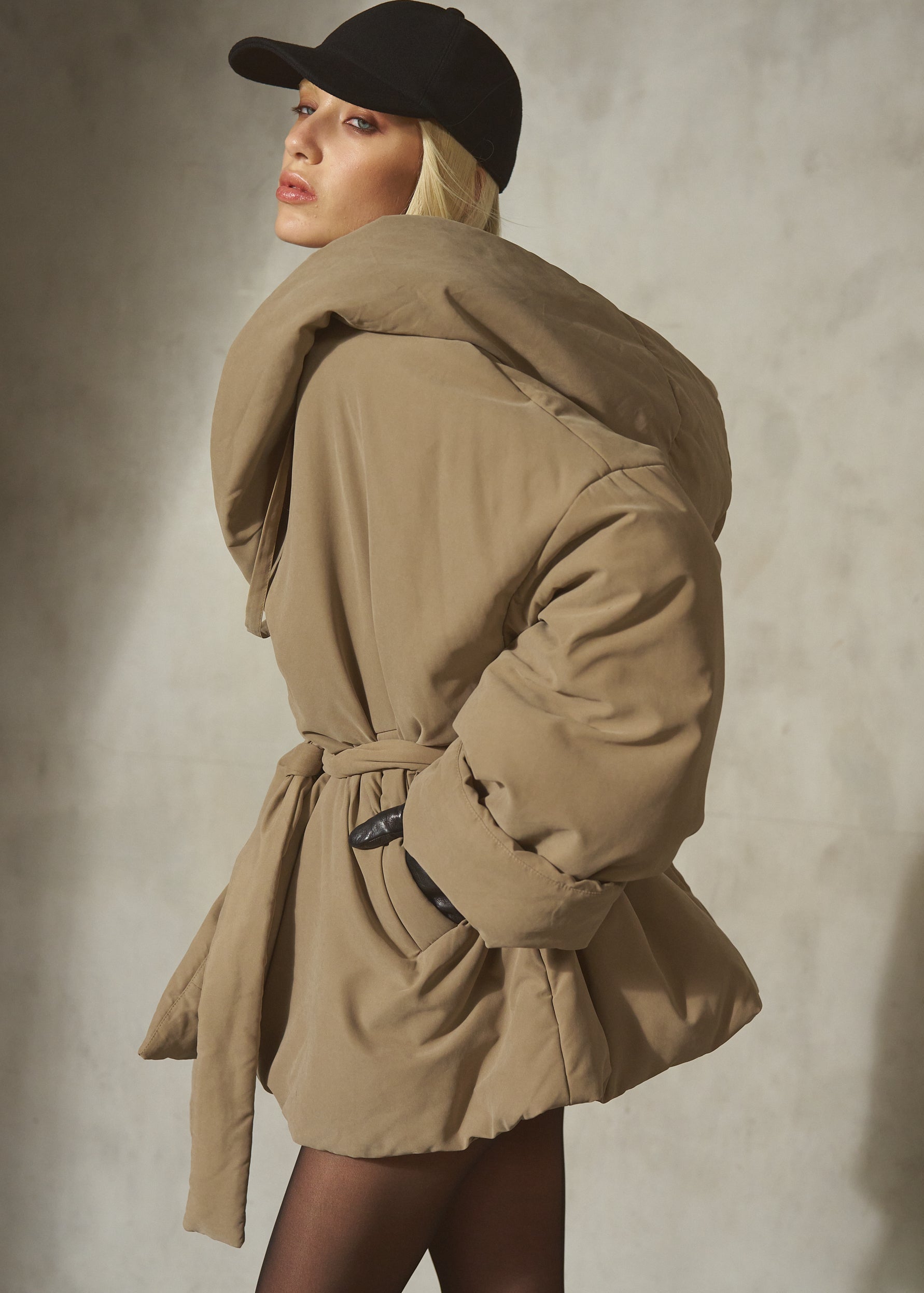 A model wearing a voluminous beige puffer coat with a matching waist tie belt, paired with a black baseball cap and sheer black tights, posing against a textured gray concrete wall.
