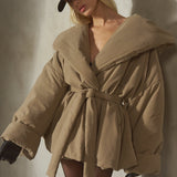 A model wearing a beige oversized belted puffer jacket, a black baseball cap, and black gloves, posing against a textured concrete wall.