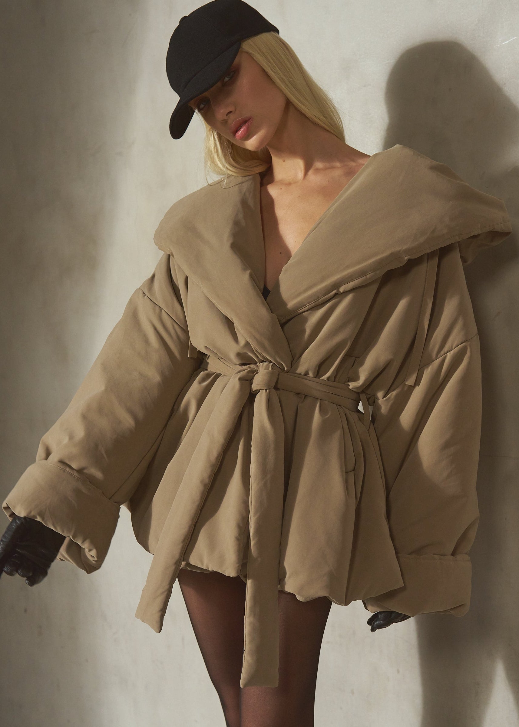A model wearing a beige oversized belted puffer jacket, a black baseball cap, and black gloves, posing against a textured concrete wall.