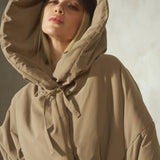 A woman wearing a beige oversized hooded jacket tied at the neck and a black cap, photographed against a soft gray studio background.
