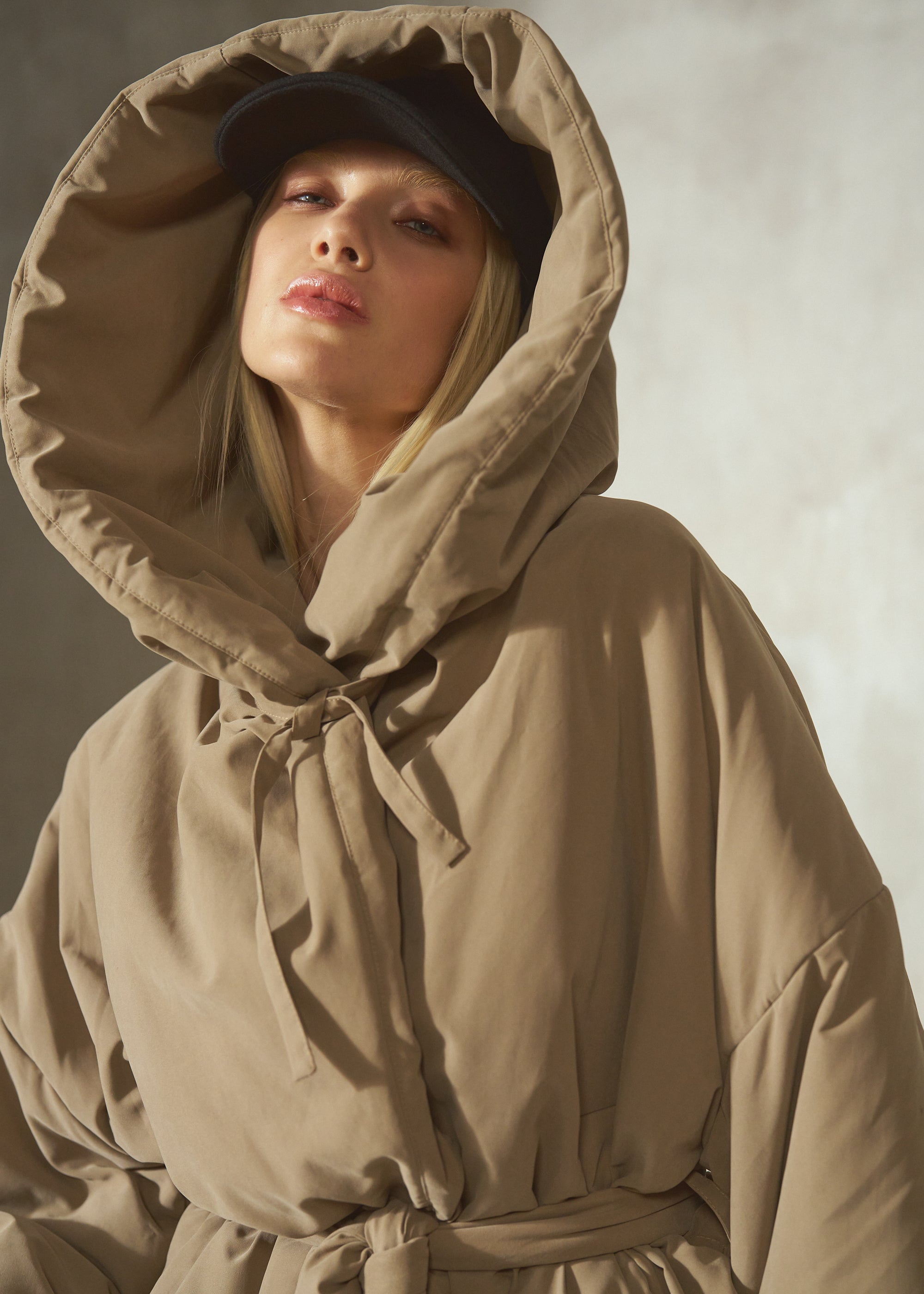 A woman wearing a beige oversized hooded jacket tied at the neck and a black cap, photographed against a soft gray studio background.
