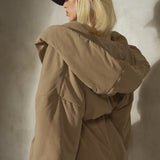 A model from behind wearing a voluminous, oversized tan hooded puffer jacket and a black wool baseball cap, standing against a textured gray concrete wall.