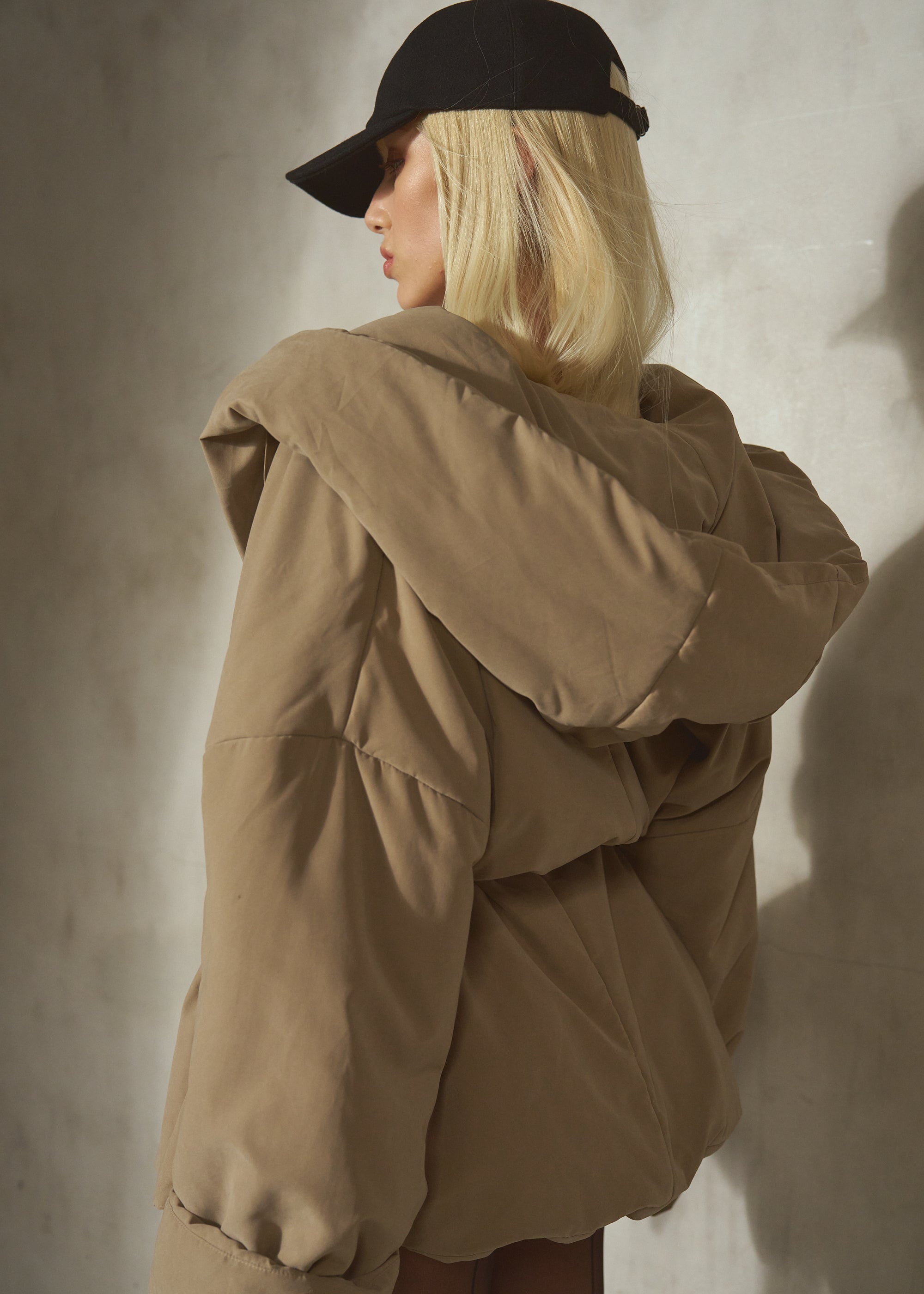 A model from behind wearing a voluminous, oversized tan hooded puffer jacket and a black wool baseball cap, standing against a textured gray concrete wall.