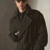 A man wearing a dark olive green cashmere trench coat over a black shirt, styled with black sunglasses and a black leather glove, standing against a textured gray wall.