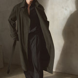 A man wearing a long dark olive cashmere trench coat over a black shirt and black trousers, standing in a minimalist concrete room with dramatic side lighting.