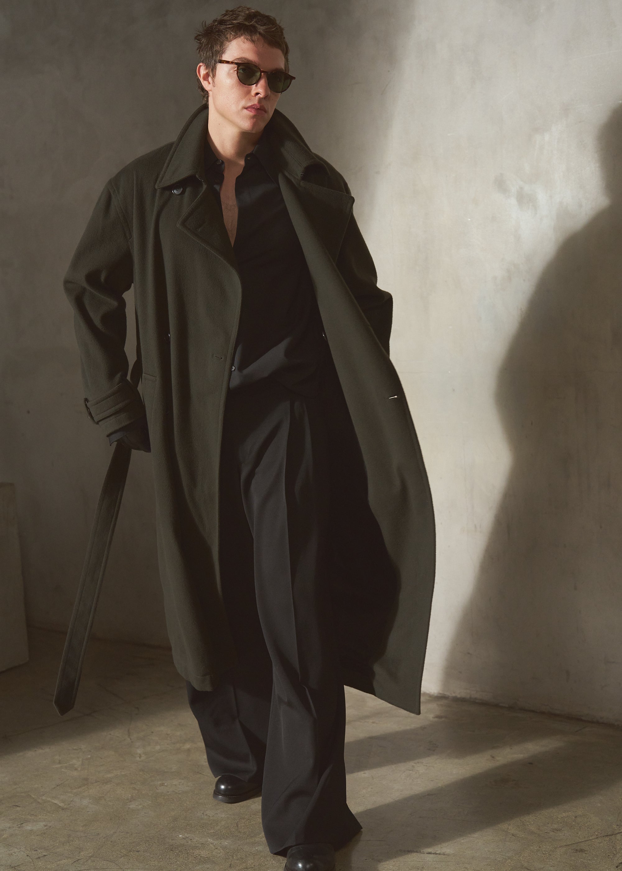 A man wearing a long dark olive cashmere trench coat over a black shirt and black trousers, standing in a minimalist concrete room with dramatic side lighting.