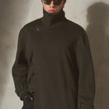A model wearing a dark olive green high-collar cashmere coat, black sunglasses, and black gloves, standing against a textured gray wall.