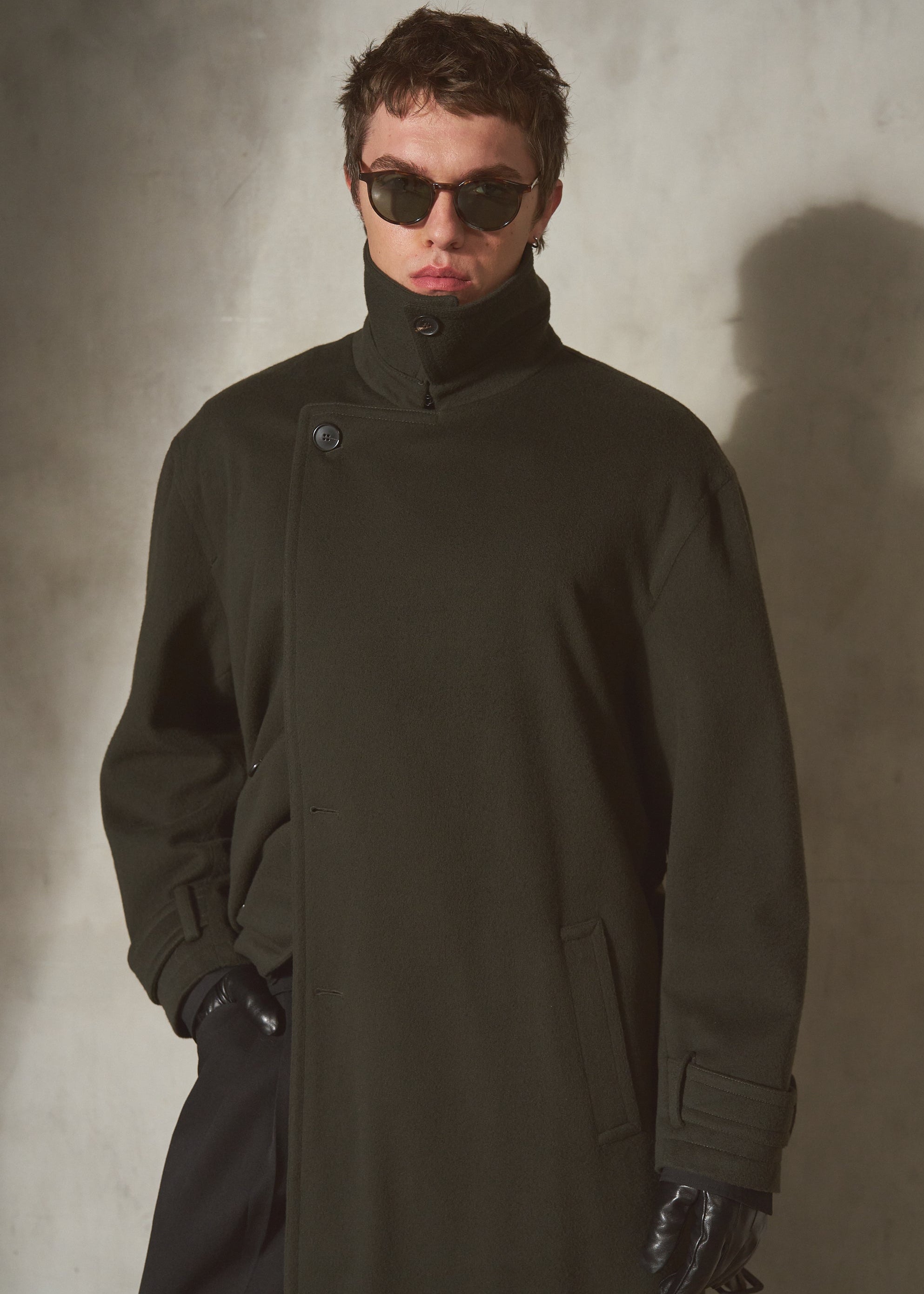 A model wearing a dark olive green high-collar cashmere coat, black sunglasses, and black gloves, standing against a textured gray wall.