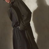 A man wearing a dark olive green trench coat over a black shirt and trousers, styled with black sunglasses, posing against a textured concrete wall with dramatic side lighting and a strong shadow.