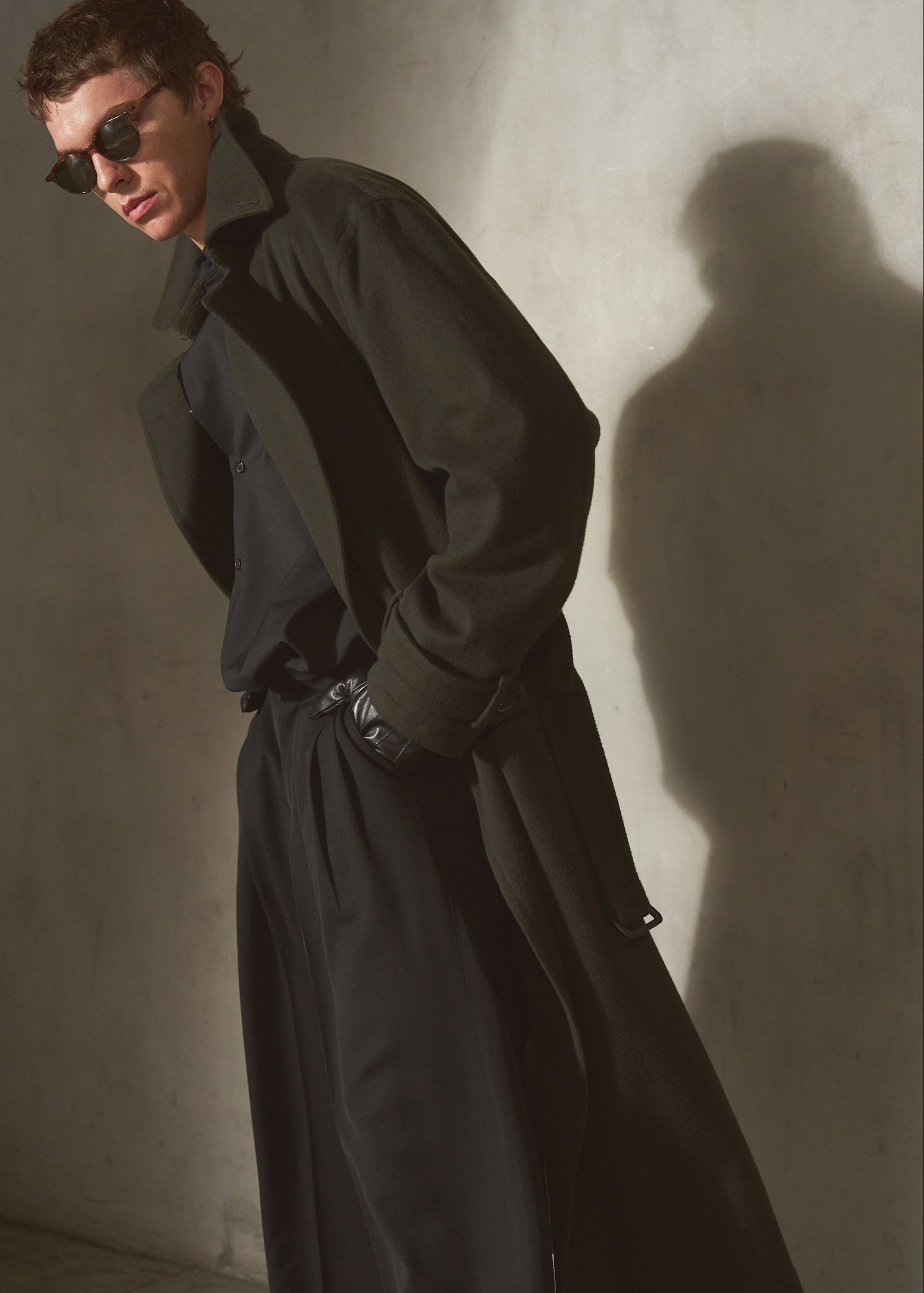 A man wearing a dark olive green trench coat over a black shirt and trousers, styled with black sunglasses, posing against a textured concrete wall with dramatic side lighting and a strong shadow.