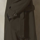 Close-up side view of a dark olive green cashmere trench coat with a matching belt, shown against a textured neutral background.