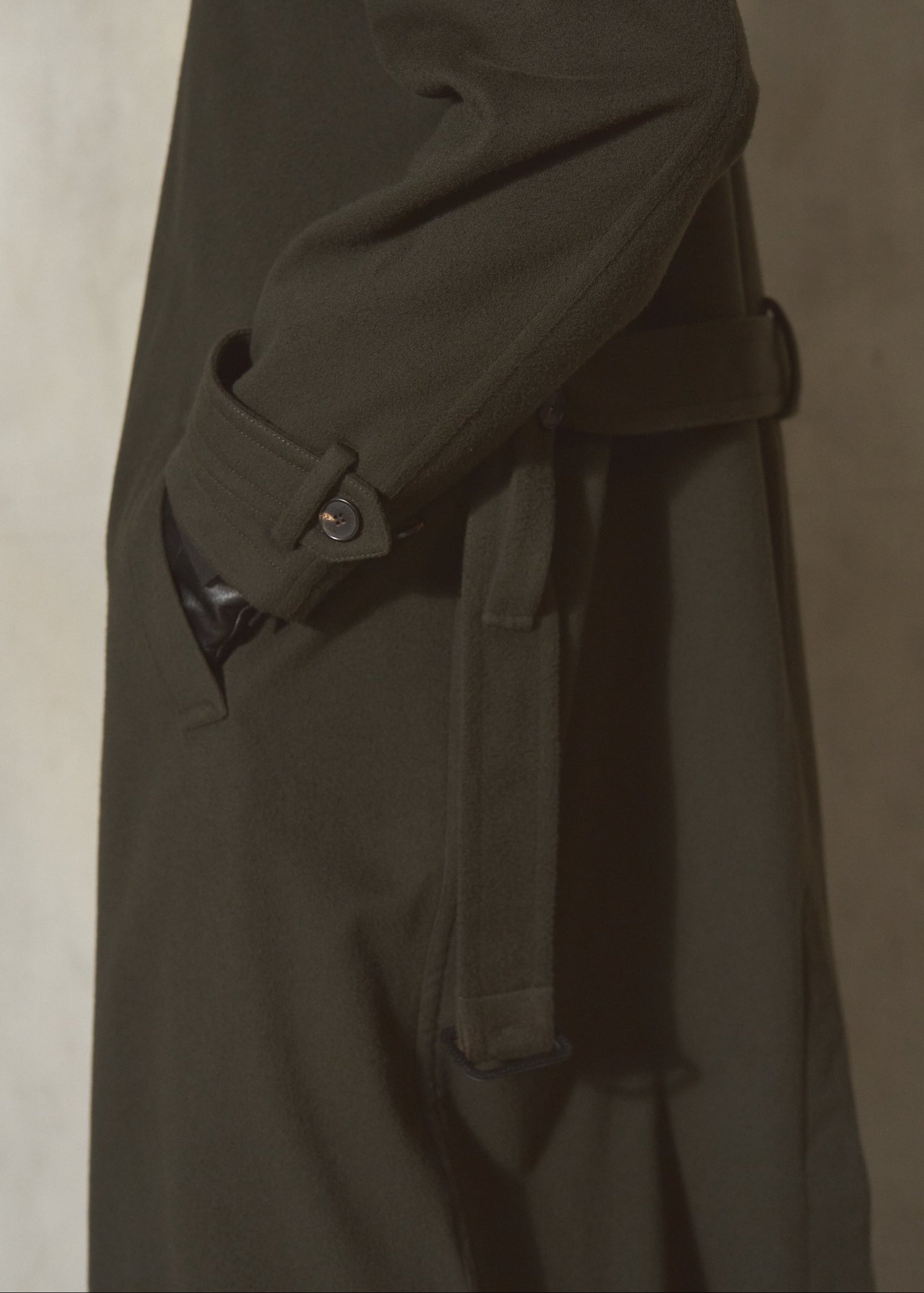 Close-up side view of a dark olive green cashmere trench coat with a matching belt, shown against a textured neutral background.