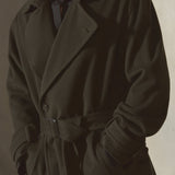 A man wearing a long dark cashmere overcoat with a matching waist belt, sunglasses, and a dark shirt, standing against a textured gray wall.