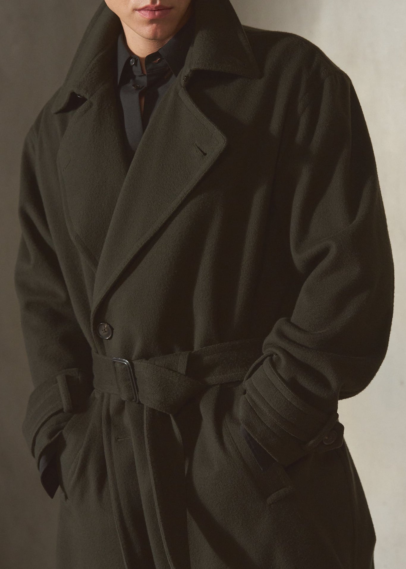 A man wearing a long dark cashmere overcoat with a matching waist belt, sunglasses, and a dark shirt, standing against a textured gray wall.