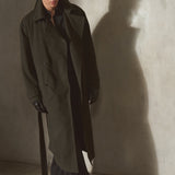 A man wearing a long dark olive cashmere trench coat, black pants, and sunglasses stands against a textured concrete wall, casting a long shadow.