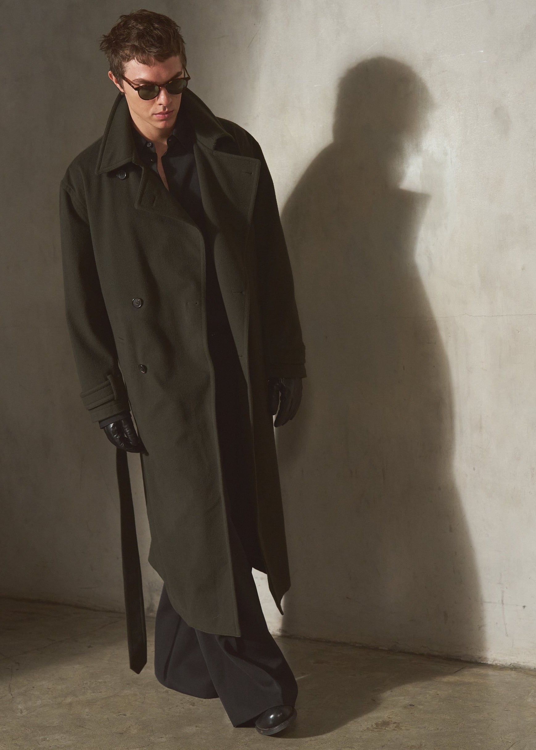 A man wearing a long dark olive cashmere trench coat, black pants, and sunglasses stands against a textured concrete wall, casting a long shadow.