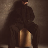 Model wearing a black cashmere wool peacoat and hat sitting on a wooden block against a textured wall.