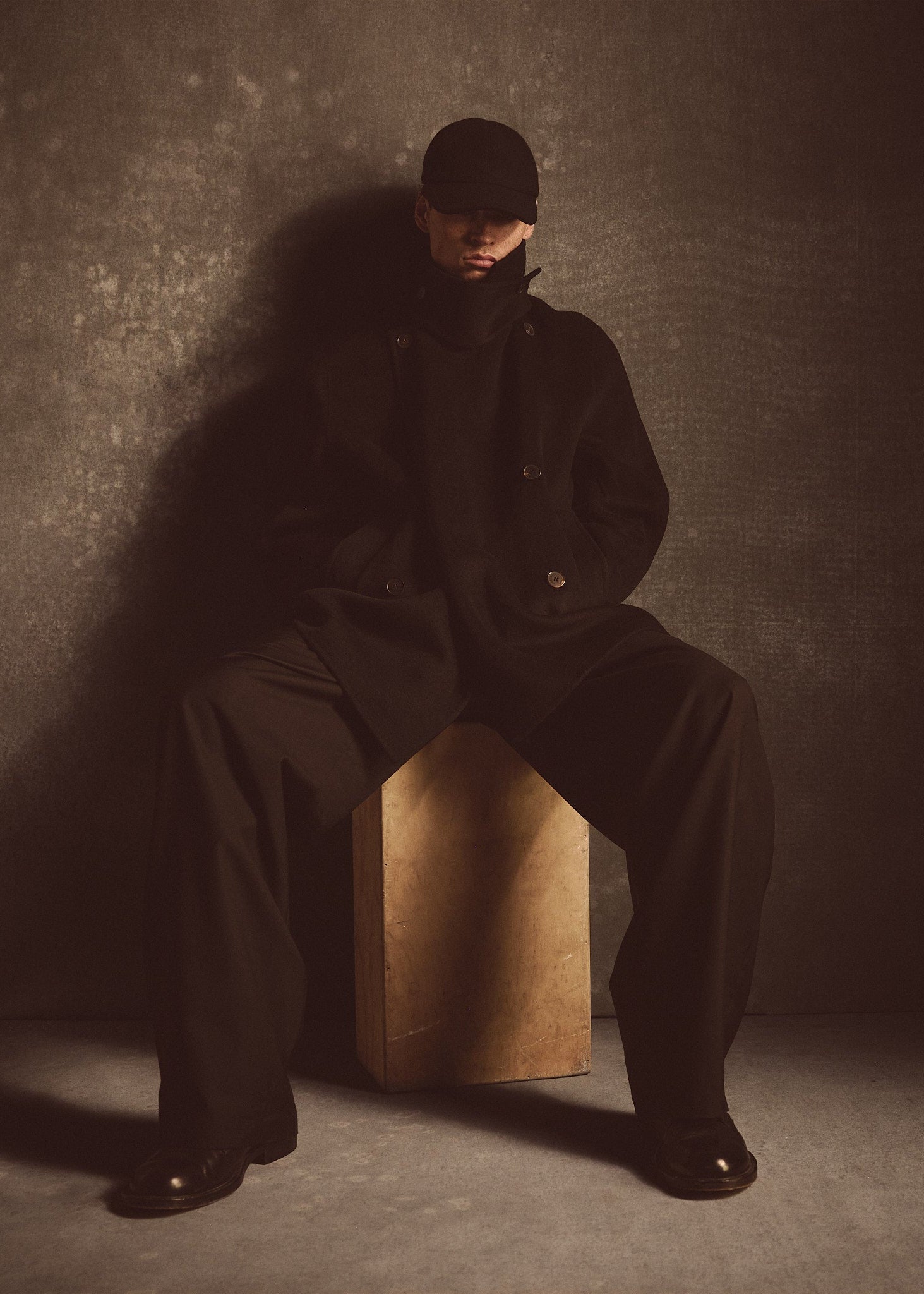 Model wearing a black cashmere wool peacoat and hat sitting on a wooden block against a textured wall.