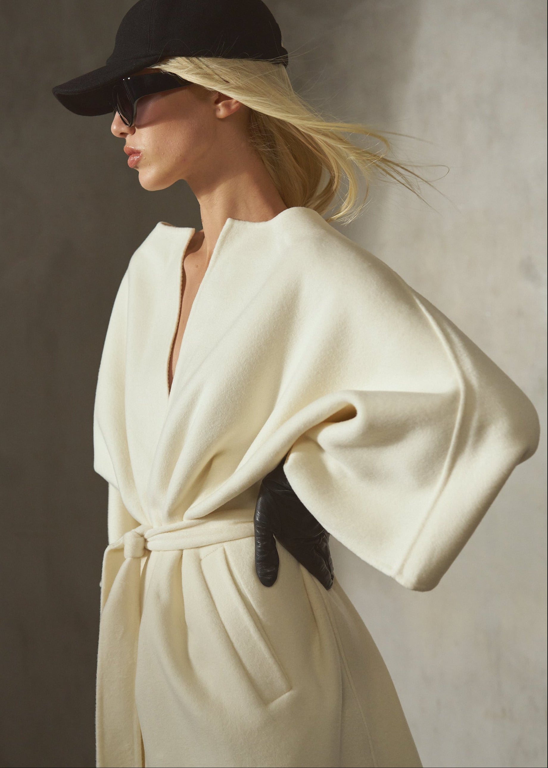 Dressed in a cashmere-wool blend, cream-colored wrap coat with wide draped sleeves and a front belt, a model wears a black cap, sunglasses, and black leather gloves, against a textured concrete wall.