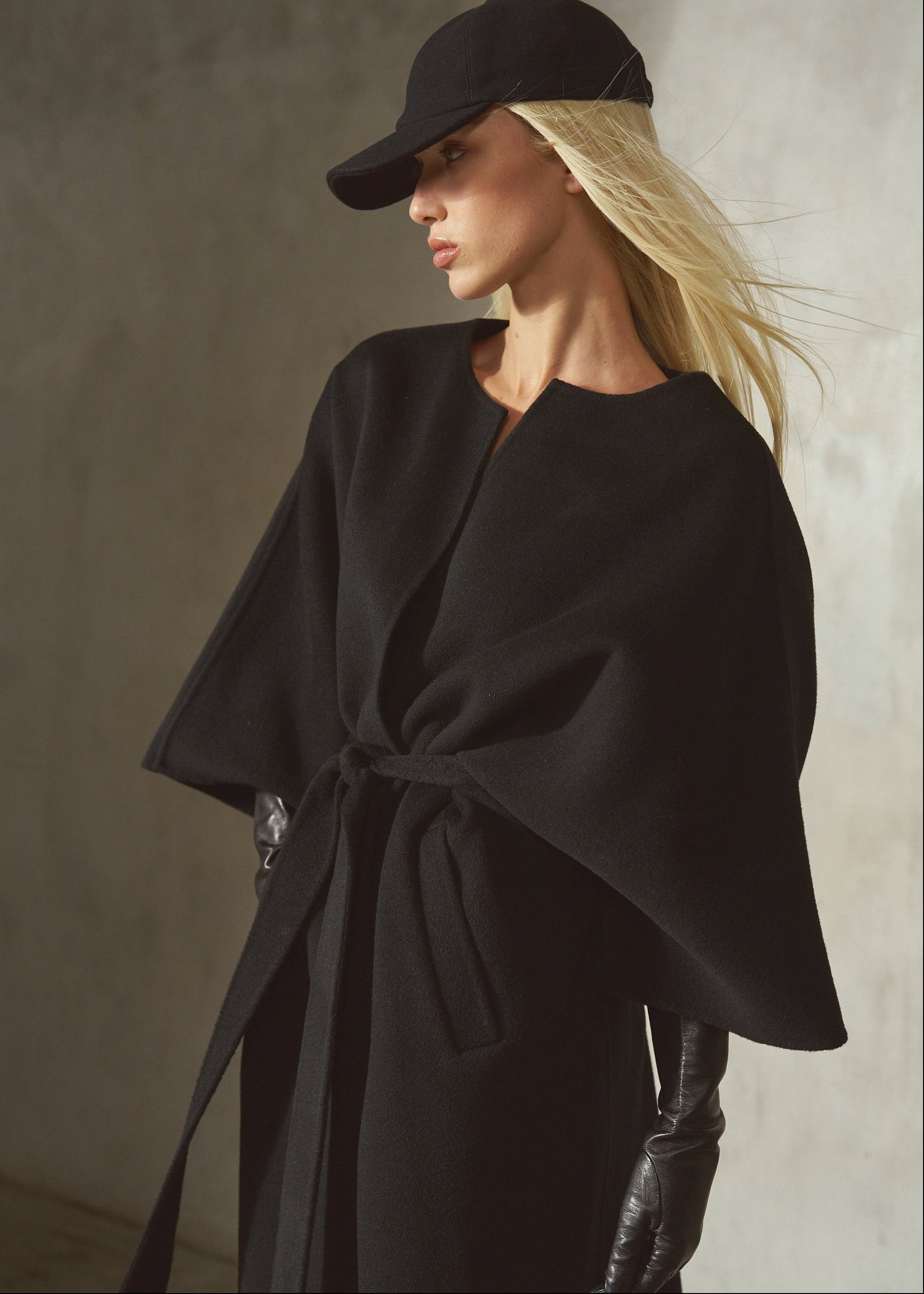 A model wears a black cashmere-wool blend wrap coat with wide draped sleeves, belted at the front, completed by a black cap and black leather gloves, against a textured concrete wall.