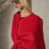 A blonde model wearing a bright red wrap-style cape coat with a matching tie belt, styled with dark sunglasses against a textured gray background.