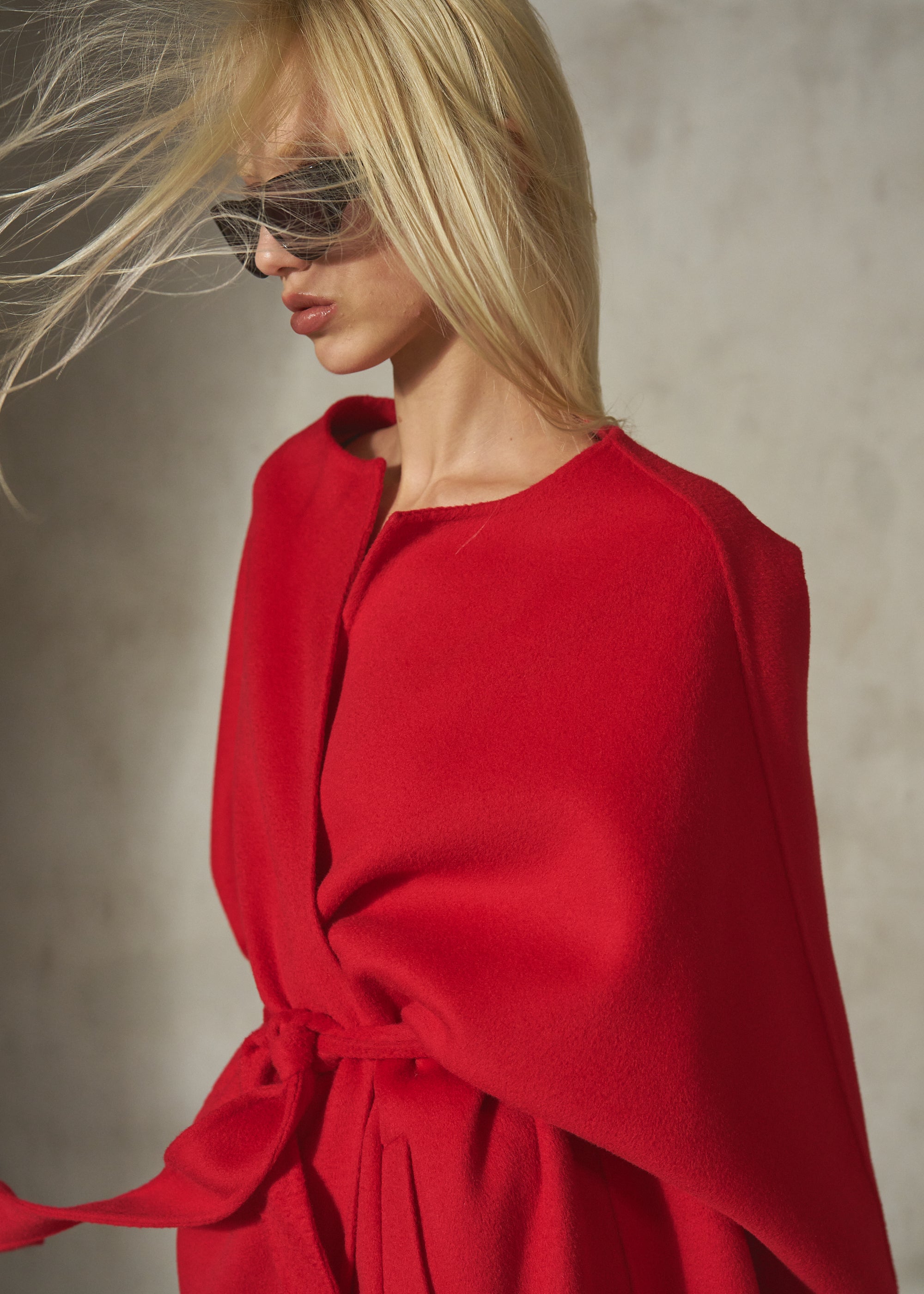 A blonde model wearing a bright red wrap-style cape coat with a matching tie belt, styled with dark sunglasses against a textured gray background.