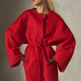 A model wearing a vibrant red oversized wrap-style coat with wide sleeves and a tie waist, styled with black sunglasses against a textured concrete wall.