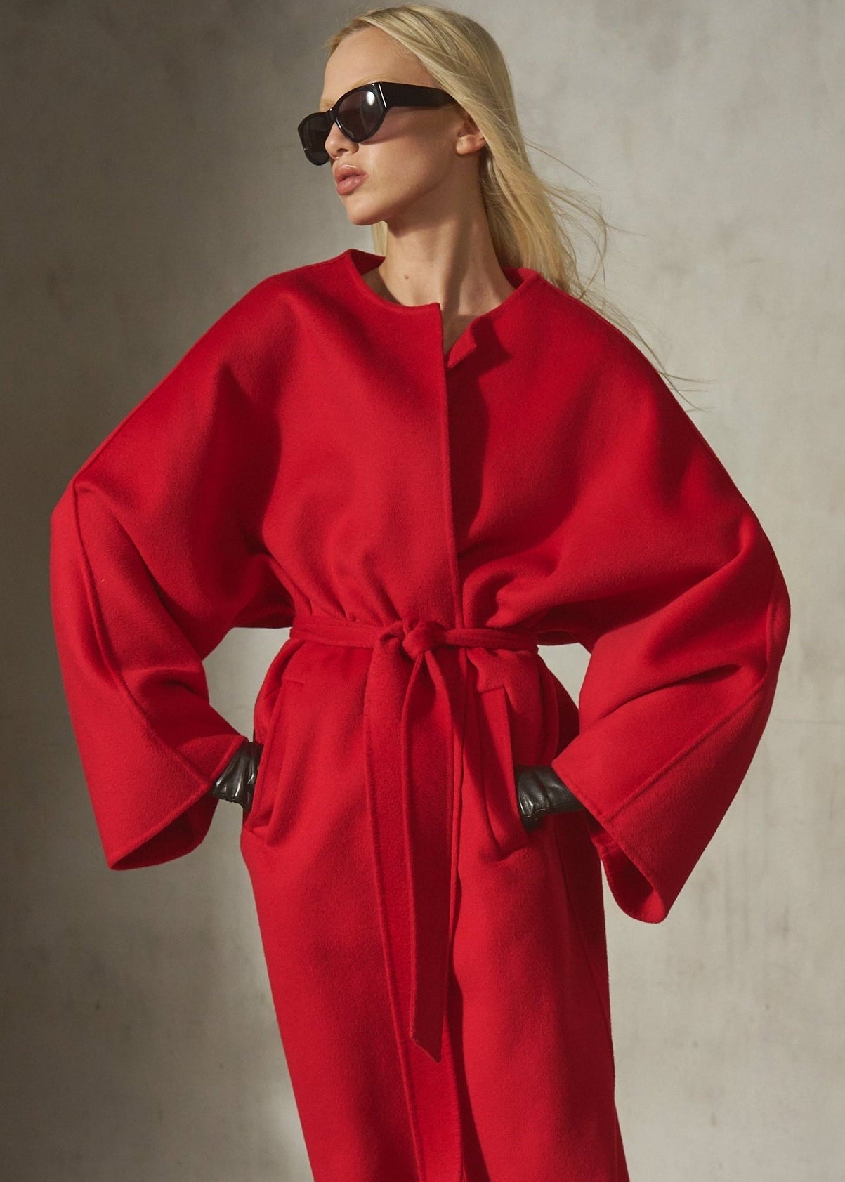A model wearing a vibrant red oversized wrap-style coat with wide sleeves and a tie waist, styled with black sunglasses against a textured concrete wall.