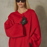 A blonde model wearing a vibrant red oversized coat, black leather gloves, and black sunglasses, posing against a textured gray concrete wall.