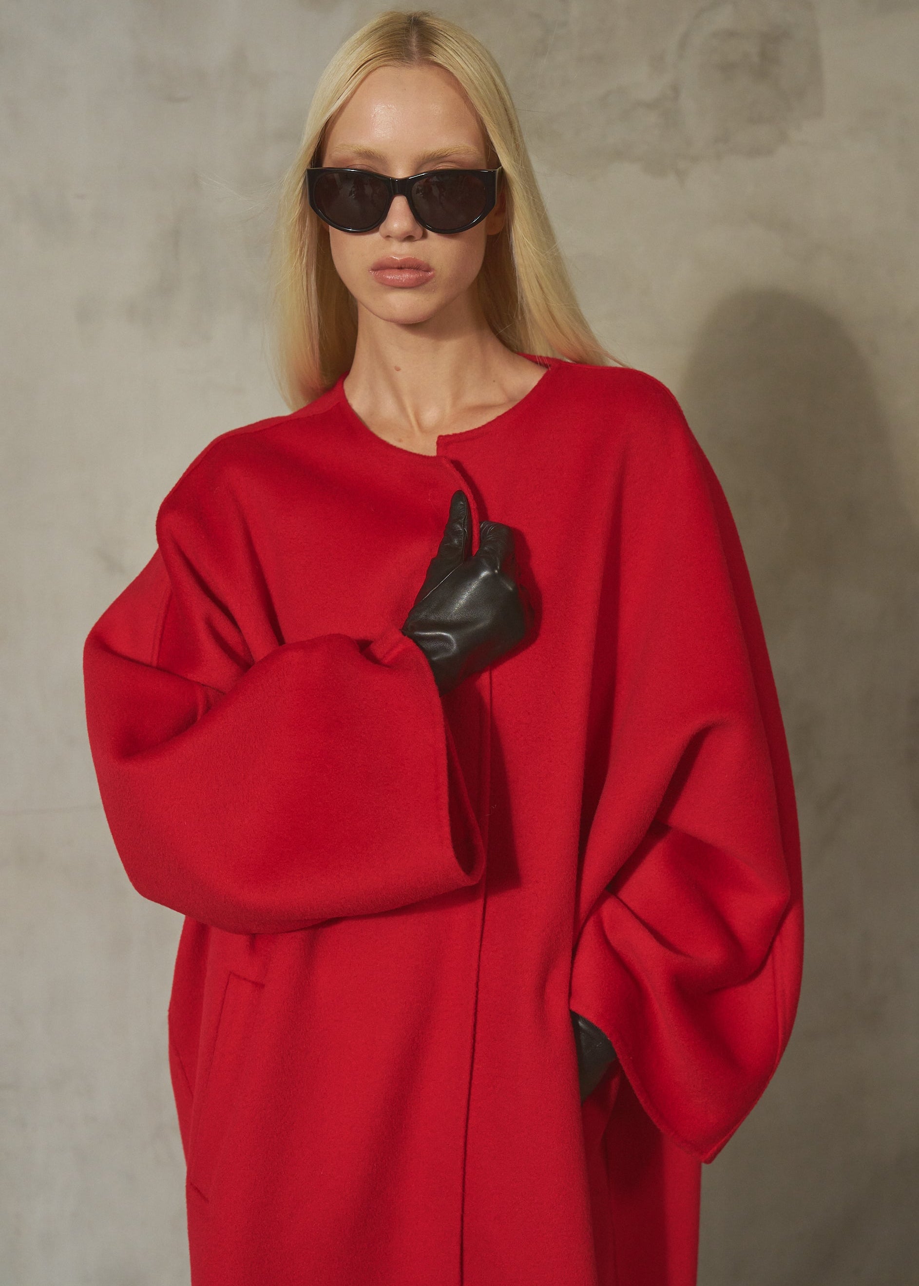 A blonde model wearing a vibrant red oversized coat, black leather gloves, and black sunglasses, posing against a textured gray concrete wall.
