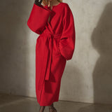 A model wearing a vibrant red belted wrap coat with dramatic oversized sleeves, styled with black gloves and dark sunglasses, standing against a textured concrete wall.