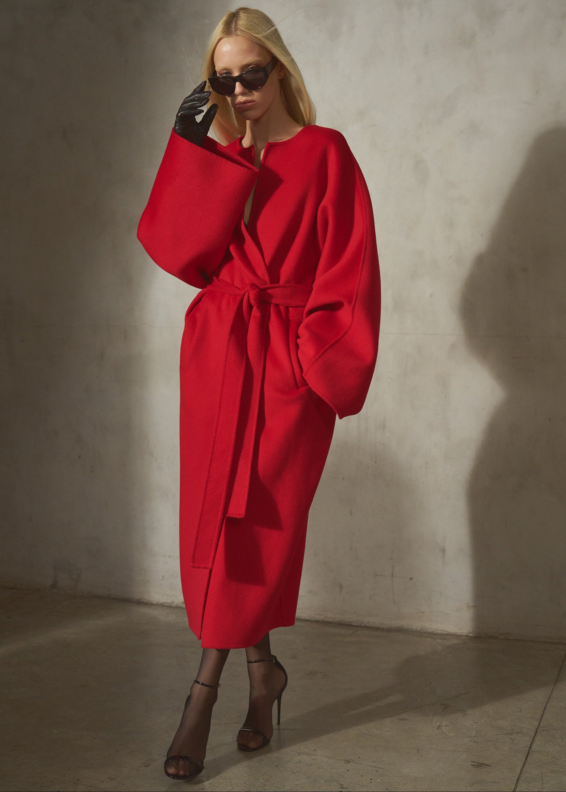 A model wearing a vibrant red belted wrap coat with dramatic oversized sleeves, styled with black gloves and dark sunglasses, standing against a textured concrete wall.