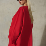 A blonde model wearing oversized black sunglasses and a voluminous bright red coat, photographed against a textured light gray wall.