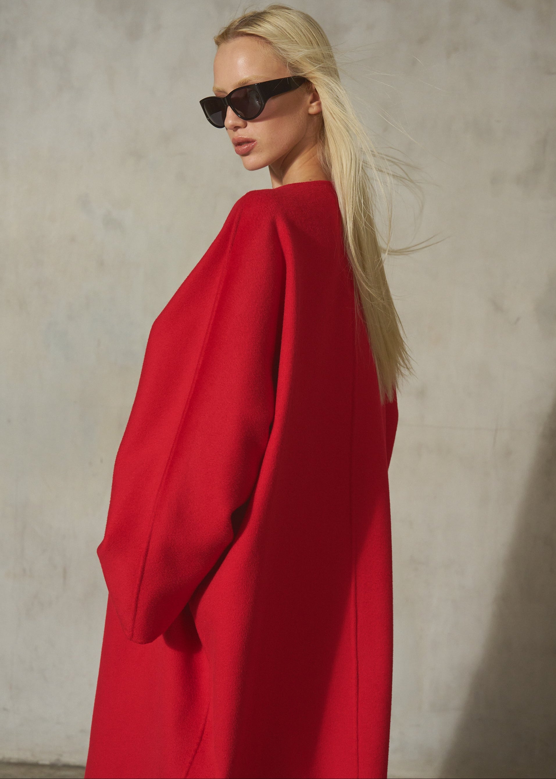 A blonde model wearing oversized black sunglasses and a voluminous bright red coat, photographed against a textured light gray wall.