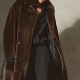 A model wearing a long brown shearling fur coat, black pants, a black shirt, and a black cap walks against a textured concrete wall with dramatic side lighting.