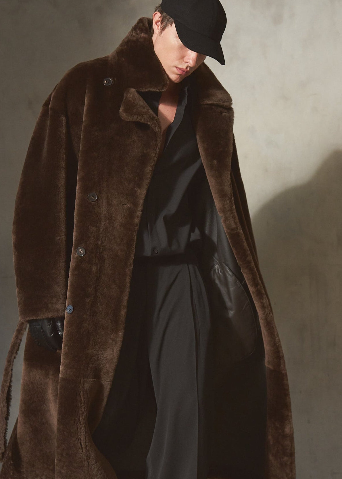 A model wearing a long brown shearling fur coat, black pants, a black shirt, and a black cap walks against a textured concrete wall with dramatic side lighting.
