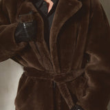 A model wearing an oversized dark brown double breasted shearling fur coat with a matching belt, black shirt, black cap, and black gloves, posed against a textured gray wall.