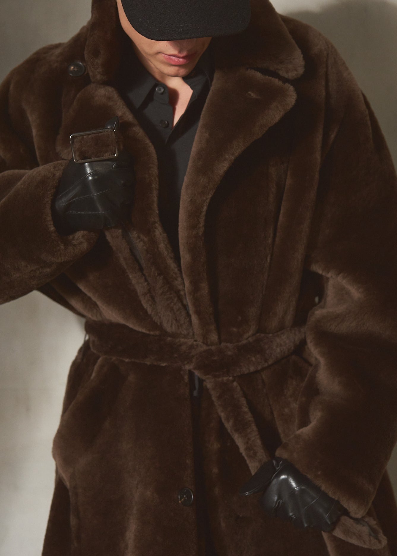 A model wearing an oversized dark brown double breasted shearling fur coat with a matching belt, black shirt, black cap, and black gloves, posed against a textured gray wall.
