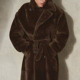 A male model wearing a long, plush dark brown double breasted shearling coat with a tie belt, black pants, and a black cap, standing against a textured off-white wall.