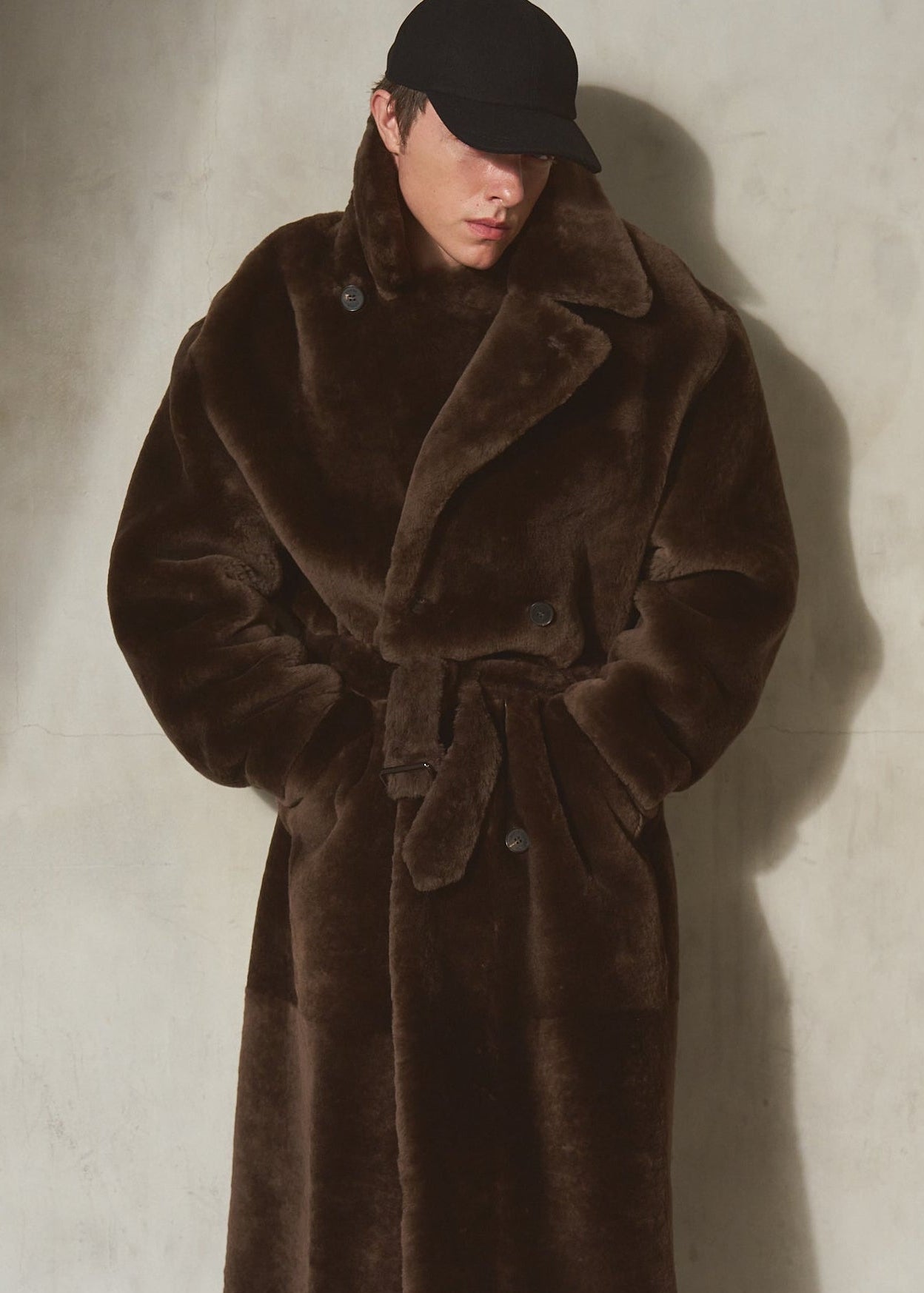 A male model wearing a long, plush dark brown double breasted shearling coat with a tie belt, black pants, and a black cap, standing against a textured off-white wall.