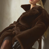 A blonde model wearing a voluminous dark brown shearling fur coat, a black baseball cap, and black tights, reclining on a wooden chair against a textured beige wall.