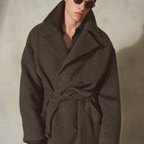 A man wearing a dark brown herringbone wool-blend belted overcoat, black leather gloves, and round sunglasses, standing against a textured light gray wall.